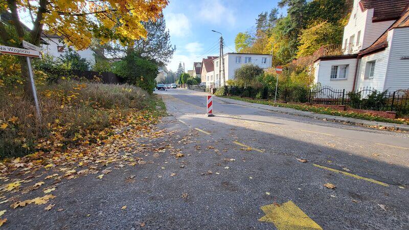 To zdjęcie przedstawia ulicę w dziennym świetle, prawdopodobnie w jesienny dzień. Widać żółte i brązowe opadłe liście, które pokrywają chodnik i krawężnik, a także żółte linie malowania drogowego. Po lewej stronie zdjęcia znajduje się znak drogowy wskazujący kierunek na "Wałbrzych". Znak jest trochę przytłoczony przez gałęzie drzewa, które wpada w kadr. Po prawej stronie widoczne są domy jednorodzinne z ogrodzeniami, a dalej, wzdłuż ulicy, zaparkowane są samochody. Środek ulicy zdaje się być wolny od ruchu. Jest to spokojna, mieszkalna okolica. Światło i cienie wskazują na porę przedpołudniową lub popołudniową. Na pierwszym planie, na środku drogi, stoi pojedynczy pachołek drogowy.