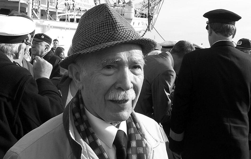 Starszy mężczyzna z wąsem siwym w kapeluszu stoi na tle nabrzeża portowego pełnego ludzi