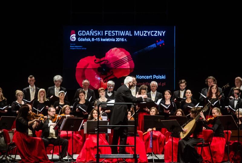 Muzycy zespołu w eleganckich strojach, kobiety ubrane w czerwone spódnice, czarne bluzki, trzymają w rękach instrumenty dawne. Na pierwszym planie dyrygent 