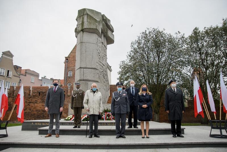 Zdjęcie przedstawia siedem osób, które stoją w dwóch rzędach i równych odstępach pod pomnikiem "Tym, co za polskość Gdańska". Twarzami skierowane są w stronę widza. Po prawej i lewej stronie znajdują się biało-czerwone flagi w naziemnych statywach. W dalszym tle widoczne są drzewa i zabudowania Głównego Miasta