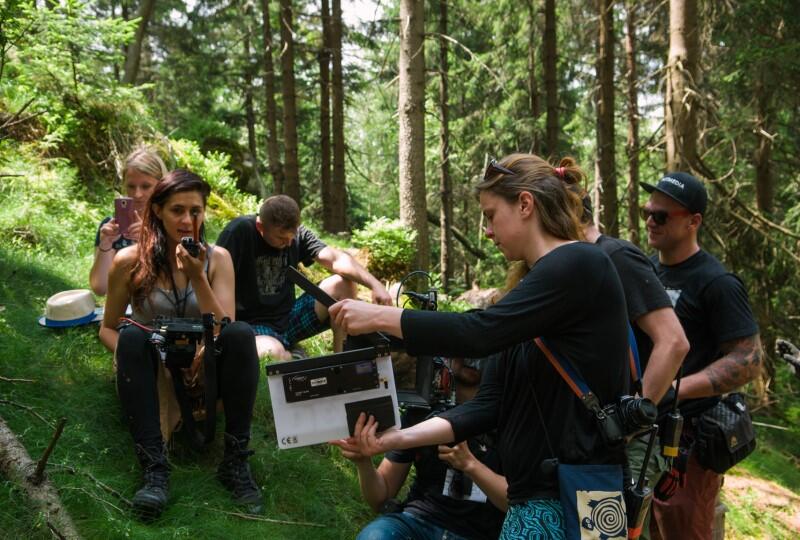 Kilka osób w lesie z kamerą i sprzętem, na pierwszym planie młoda kobieta z długimi ciemnymi włosami, ma apart fotograficzny przewieszony przez ramię, w ręku trzyma filmowy klaps