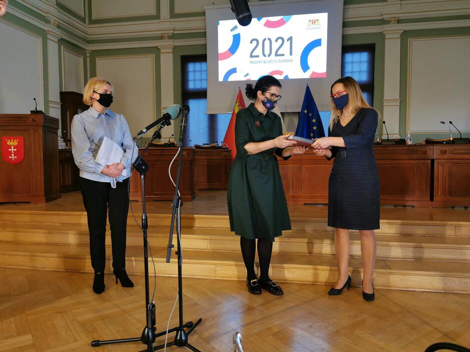 Na zdjęciu widać Prezydent Aleksandrę Dulkiewicz ( w środku), która przekazuje papierową wersję projektu budżetu Gdańska na 2021 rok przewodniczącej Rady Miasta Agnieszce Owczarczak. W wydarzeniu uczestniczyła także Emilia Kosińska, zastępca Skarbnik Miasta (po lewej)