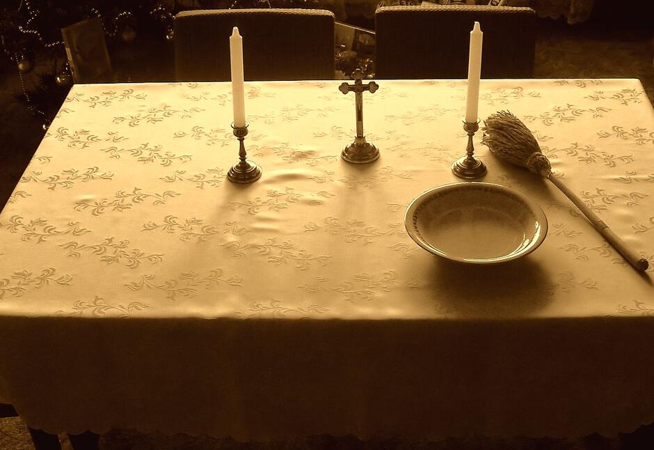 stół w pokoju na nim biały obrus, kropidło, krzyż, woda święcona i świece, w tle choinka