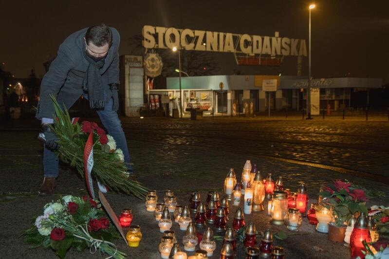 mężczyzna w płaszczu składa wiązankę kwiatów przy tablicy, stoją świece