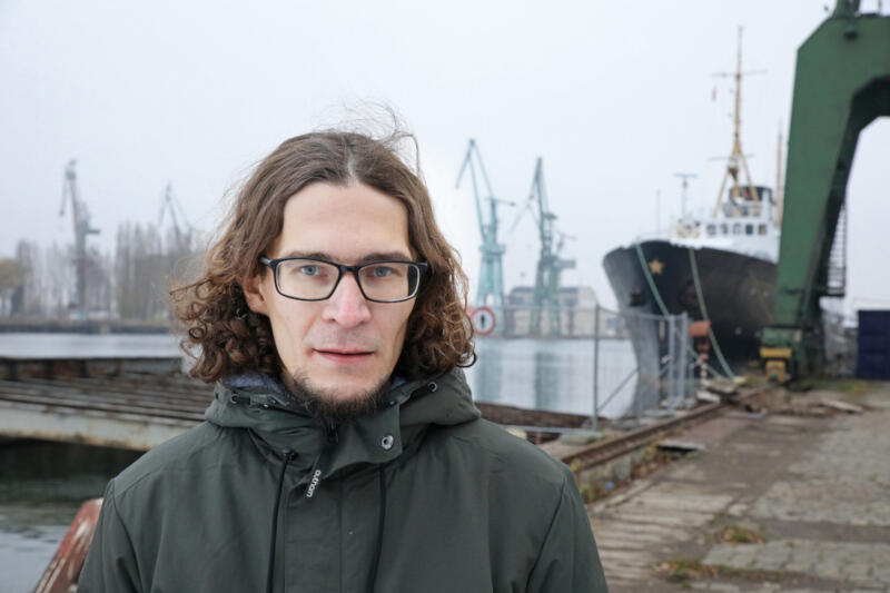 Na zdjęciu portretowym Ilia Pyzhin, młody mężczyzna, szatyn z bródką, nieco dłuższe niż za uszy włosy, okulary, patrzy w obiektyw. Za nim dźwigi stoczniowe. Ma na sobie ciemno-zieloną zimową kurtkę