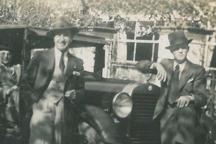 Czarno-białe zdjęcie przedstawiające dwóch mężczyzn pozujących na tle samochodu z epoki