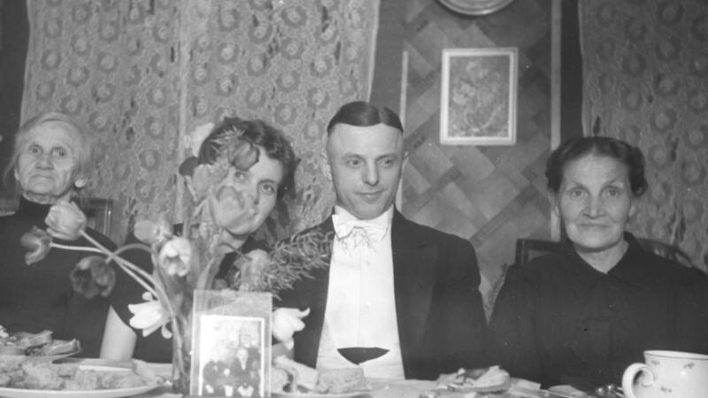 czarno-białe zdjęcie, cztery osoby siedzą przy zastawionym stole, na którym stoi m.in. bukiet kwiatów i fotografia w ramce. Trzy kobiety i mężczyzna - drugi od prawej, elegancko ubrany, w białej koszuli i muszce, uczesany na gładko