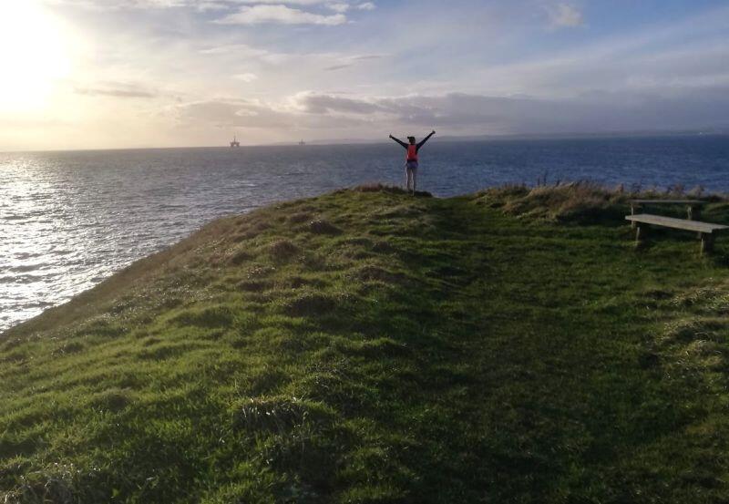 piękny widok morza i zachodzącego słońca widziany ze skał Szkocji, na skale stoi młoda kobieta w sportowym stroju i unosi ręce
