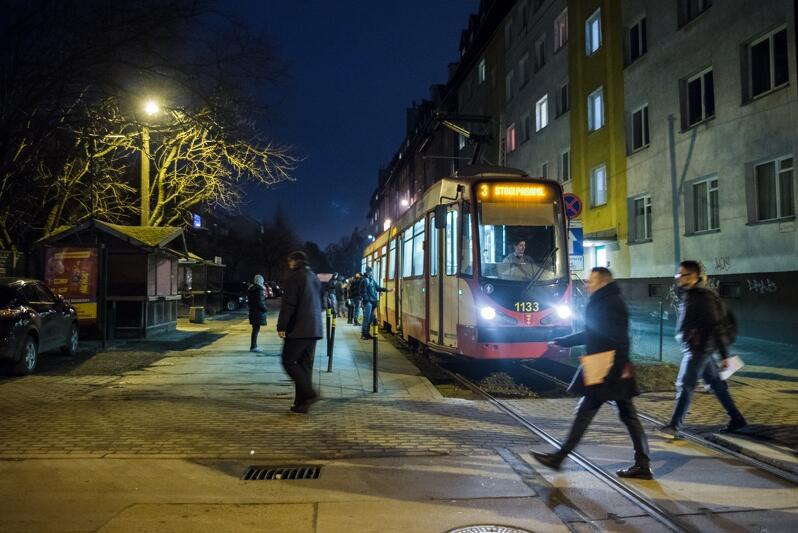 zdjęcie wykonane wieczorową porą, na pierwszym planie dwóch mężczyzn przechodzi przez tory tramwajowe, obok nich stoi oświetlony tramwaj w kolorach biało-czerwonym, po prawej stronie widac kilkupiętrowy szary budynek mieszkalny