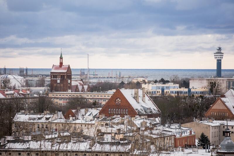 zdjęcie wykonane z dużej wysokości, widać na nim dachy budynków, głównie kamienic, oprószone śniegiem, wyróżnia się wieża kościelna, a także wieża w porcie morskim, w tle fragment zatoki