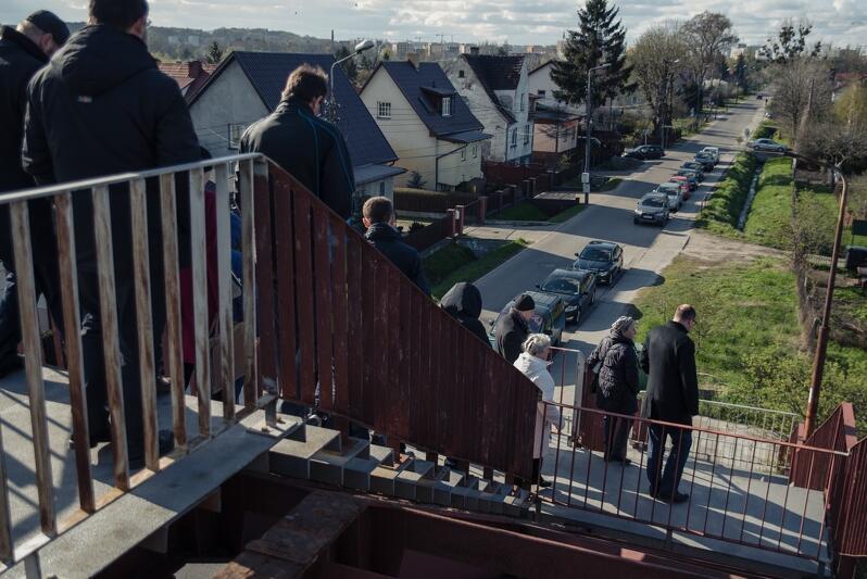 na zdjęciu widać grupę osób w jesiennych ubraniach, schodzących po schodach otoczonych zniszczonymi zardzewiałymi barierkami, w tle po lewej widać ustawione w jednym szeregu domki jednorodzinne ze spadzistymi czarnymi dachami, widać też fragment jezdni