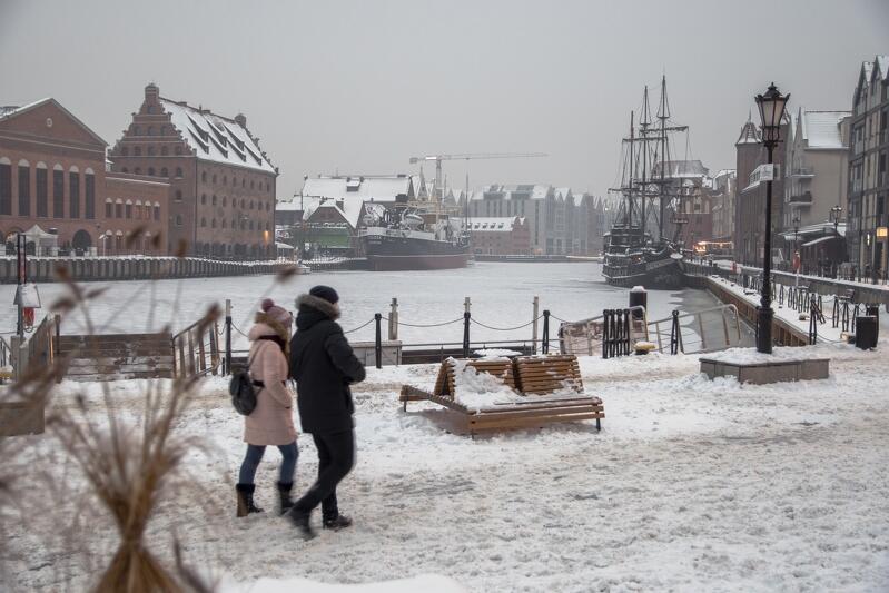 zdjęcie gdańskiego pobrzeża, widać parę osób spacerujących po zaśnieżonym chodniku, stoją tam ławki i latarnie, widać też fragment rzeki Motławy, przy jednym brzegu cumuje statek Sołdek, przy drugim - po prawej - zabytkowy statek turystyczny, po obu stronach widać zabytkowe budynki Gdańska