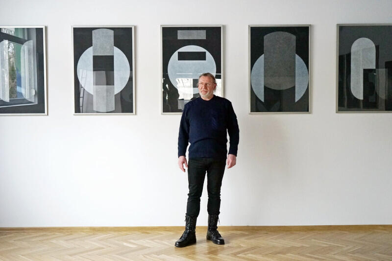 Stanisław Olesiejuk, artysta pozuje w galerii - białe ściany, jasne, drewniane podłogi - na tle swoich prac. Kilka czarno-białych rysunków wisi na ścianie. Artysta jest ubrany na czarno