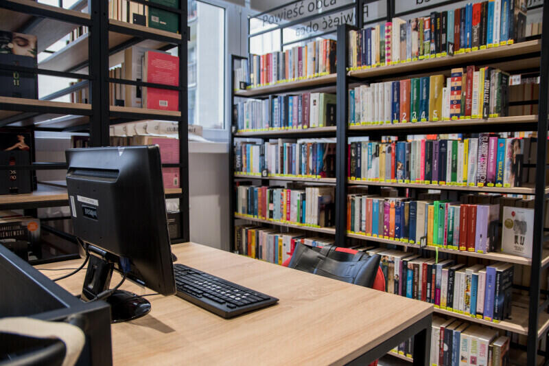 Wnętrze biblioteki, po lewej stronie biurko z płaskim czarnym ekranem komputera, w tle i po prawej stronie metalowo-drewniane regały z książkami