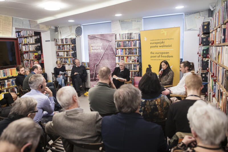 17 osób siedzi w bibliotece, w tle regały z książkami i dwa duże banery, jeden z napisem Instytut Kultury Miejskeij, drugi z napisem Europejski poeta wolności