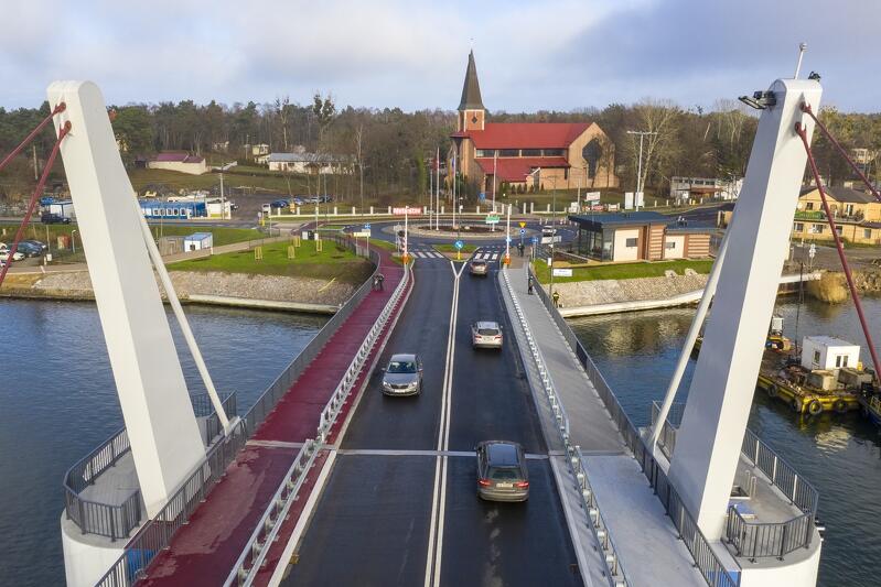 zdjęcie wykonane z drona, widać fragment mostu zwodzonego po którym przejeżdża kilka samochodów osobowych, za mostem widać rondo, a za nim kościół, po obu stronach mostu widać wody rzeki Martwej Wisły, 