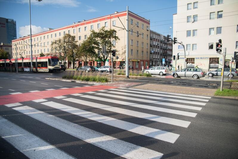 na pierwszym planie widać przejście dla pieszych wyznaczone białymi pasami, jest też czerwona droga dla rowerów, w tle dwa budynki wzdłuż ulicy, jest tez fragment jezdni po której jadą samochody, po lewej widać jadący biało-czerwony tramwaj.