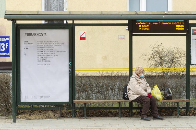 przystanek autobusowy z plakatem, na ławce siedzi starsza kobieta w maseczce, skierowana w bok, obok niej leżą na ławce siatki z zakupami