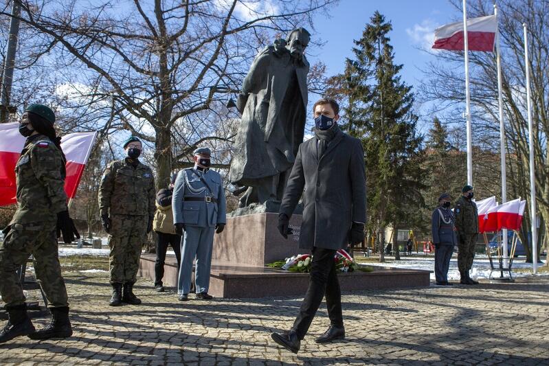 Kilka postaci pod pomnikiem Piłsudskiego na Strzyży. Dwie z nich odchodzą po złożeniu kwiatów. Centralnie widoczny jest wysoki młody mężczyzna, Piotr Grzelak. W tle widzimy drzewa parku. Po obu stronach pomnika stoją po dwie osoby jako straż honorowa, a także znajdujące się w statywach biało-czerwone flagi