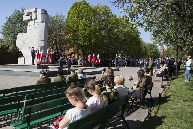 Po lewej widoczny jest pomnik Tym, co za polskość Gdańska. Resztę ilustracji zajmuje przestrzeń skweru. Na pierwszym planie są ławki, na których siedzi kilka młodych osób. Przy pomniku trwa uroczystość patriotyczna, są biało-czerwone flagi, warta honorowa i tłum ludzi