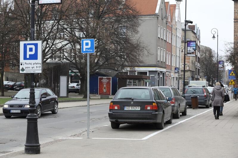 Widok ulicy w Śródmieściu Gdańska. Na miejscach parkingowych, przy poboczach chodników, zaparkowane są samochody - jeden za drugim