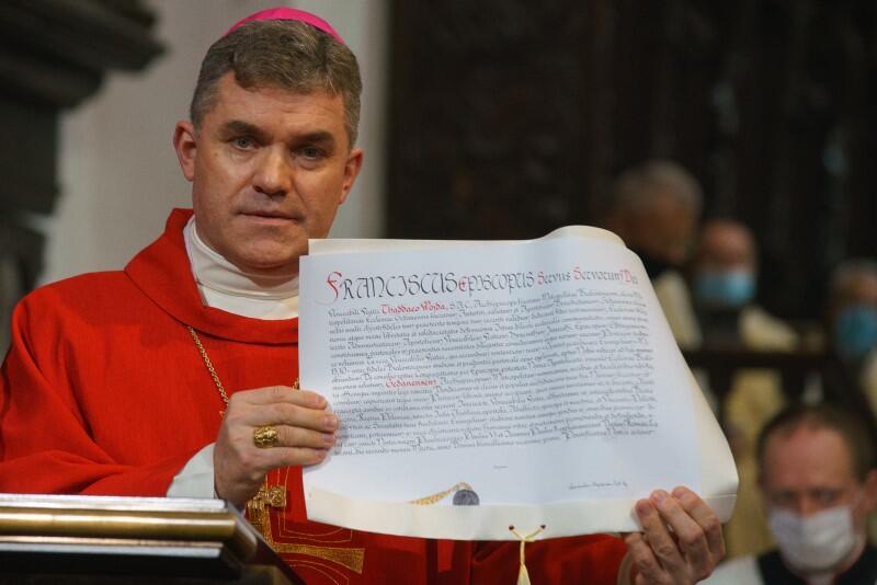 Mężczyzna w średnim wieku, w szatach biskupich rozpościera w rękach duży dokument z pieczęcią papieską i prezentuje go publiczności