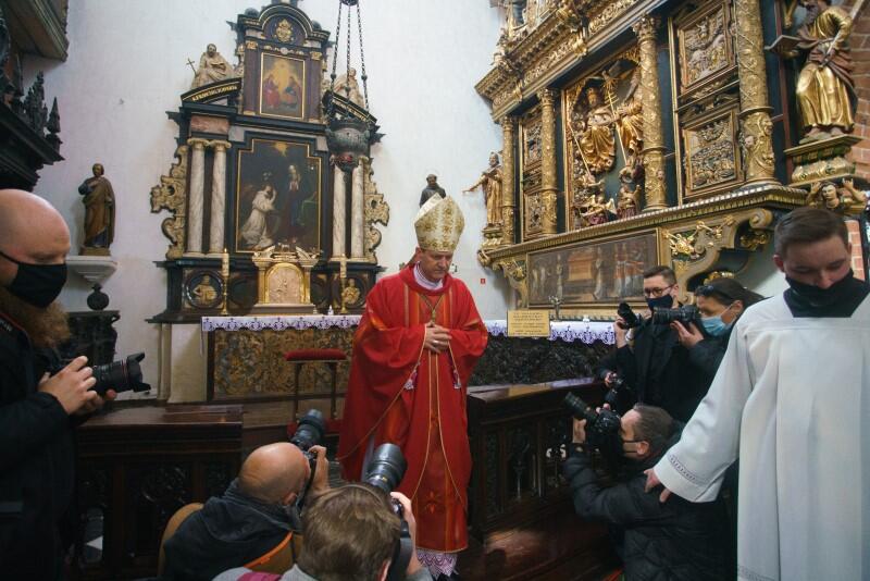 Arcybiskup Wojda stoi w prezbiterium  Katedry Oliwskiej, z dłońmi splecionymi na piersi. Otaczają go fotoreporterzy robiący zdjęcia