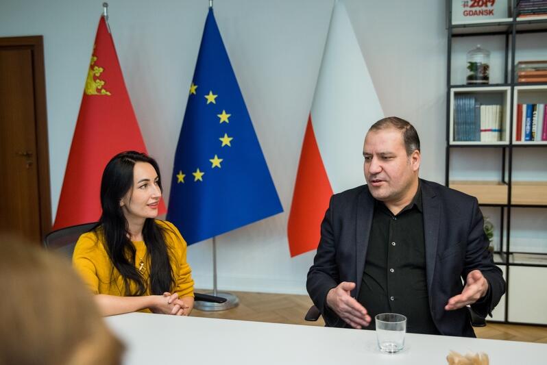 mężczyzna siedzi za stołem, gestykuluje podczas rozmowy, za nim widnieją trzy flagi: Polski Gdańska i Unii Europejskiej
