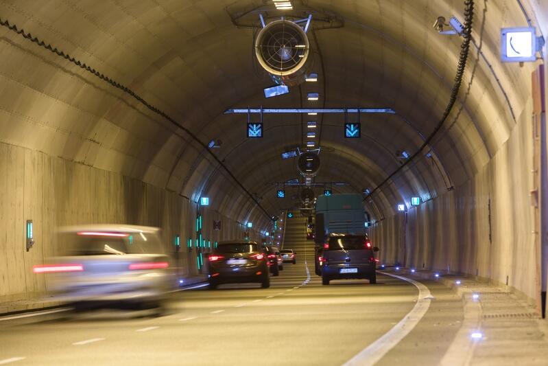 Wnętrze tunelu pokazane zza przedniej szyby jadącego samochodu. Widać auta, które są z przodu - wszystkie jadą w tym samym kierunku, po dwóch pasach jezdni. Sklepienie tunelu jest półkoliste. W szczytowym punkcie sklepienia podwieszony jest duży wentylator do usuwania spalin z tunelu