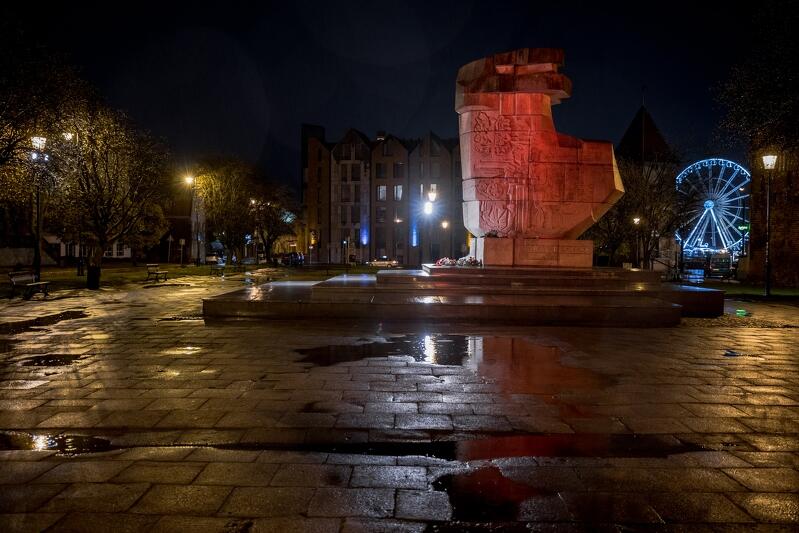 Ciemno, plac z kałużami wody. Na placu - trochę w oddali - pomnik Tym, co za Polskość Gdańska oświetlony na czerwono 