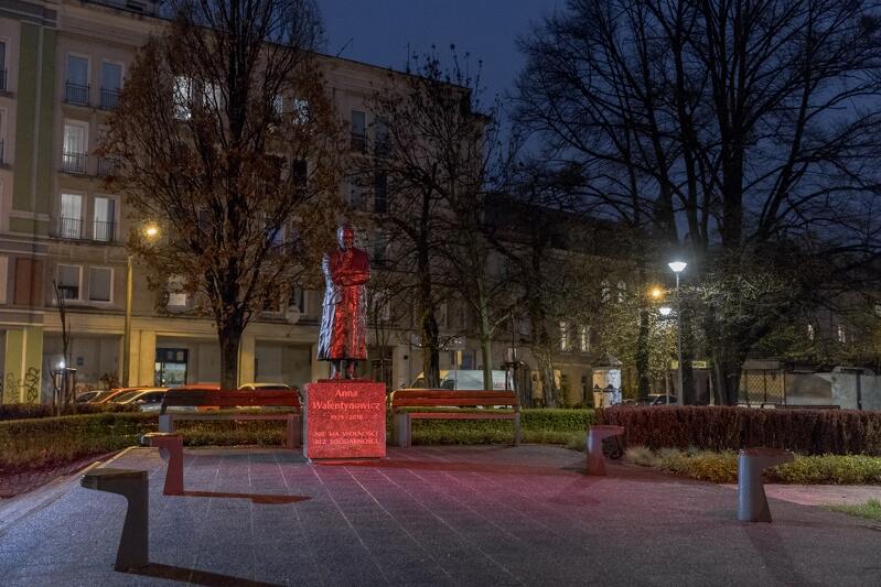Wieczór, prawie ciemno. Plac chodnikowy, na nim pomnik Anny Walentynowicz podświetlony na czerwono. Za nim dwa drzewa, jeszcze dalej budynki wielopiętrowe