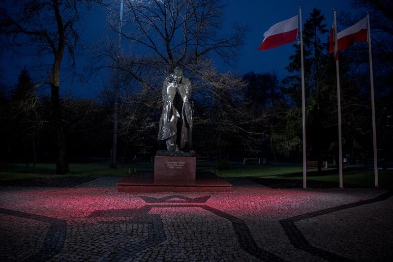 Wieczór, prawie ciemno. Plac w bruku, na nim pomnik marszałka Józefa Piłsudskiego podświetlony: cokół na czerwono, Marszałek na biało. Za nim zieleń - trawnik, krzewy, drzewa. Obok trzy maszty, na nich biało-czerwone flagi