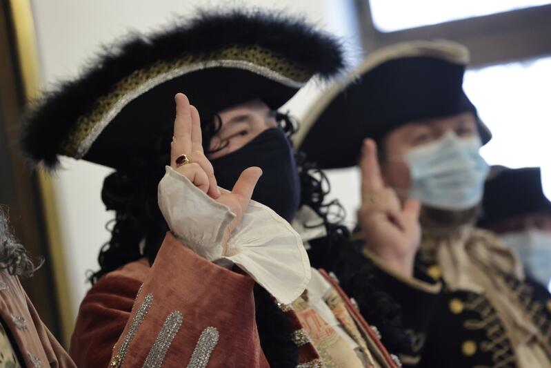 Dwóch mężczyzn ubranych w stroje z XVI wieku, mają podniesione prawe dłonie, trzy palce wyciągnięte ku górze. Na twarzach mają maseczki ochronne 