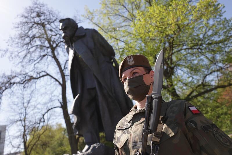 górujący pomnik w tle - postać mężczyzny, na pierwszym palnie żołnierka z karabinek i w masce 