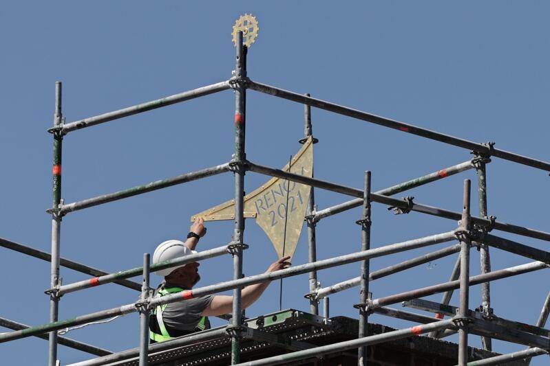 pokryta złotem chorągiew ustawiana jest przez robotnika w kasku na szczycie dachu 