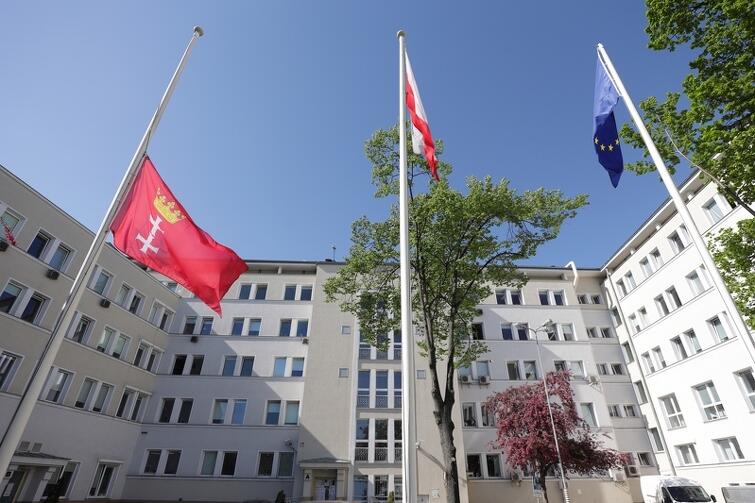 Trzy flagi na masztach otoczonych białymi budynkami. Jedna z nich - czerwona flaga Gdańska - opuszczona jest do połowy masztu