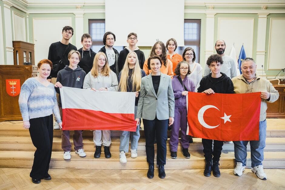 Grupowe zdjęcie około 20 osób w sali obrad Rady Miasta Gdańska. Głównie młodzież szkolna. Dwie osoby na przedzie trzymają rozpostarte flagi państwowe. Po lewej jest flaga polska, po prawej - turecka 