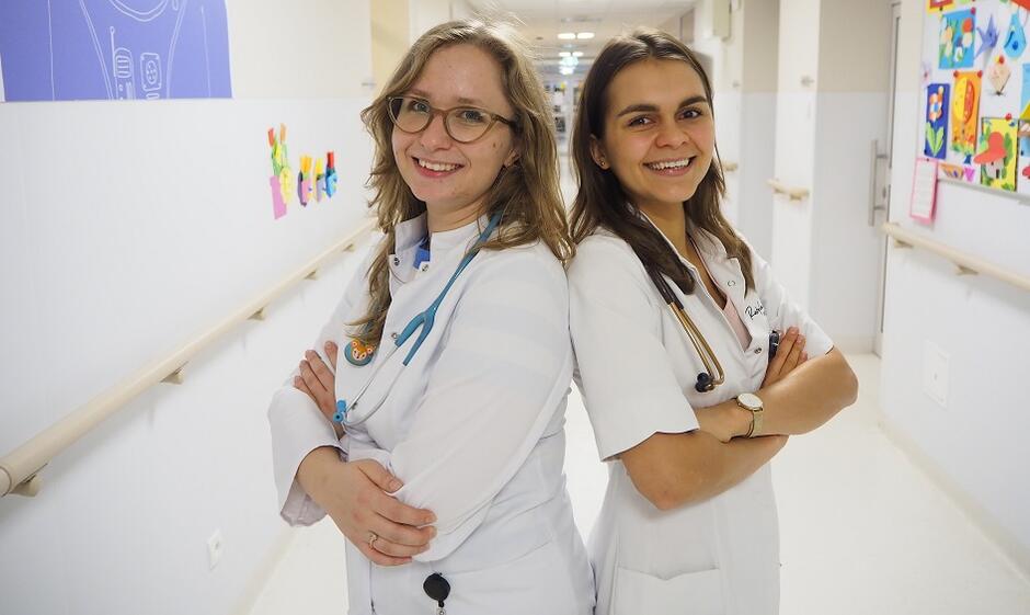dwie młode kobiety w lekarskich fartuchach na szpitalnym korytarzu, zwrócone do siebie plecami, uśmiechnięte