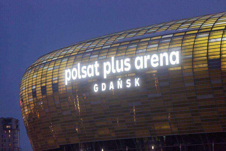 Zrobione w ciemności zdjęcie. Widać kontury stadionu i świecący w ciemności na biało napis Polsat Plus Arena Gdańsk