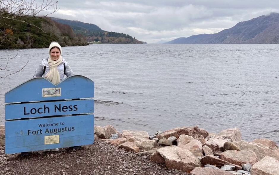 młoda kobieta w czapce na tle jeziora stoi w chłodny dzień przy tabliczce z napisem Loch Ness