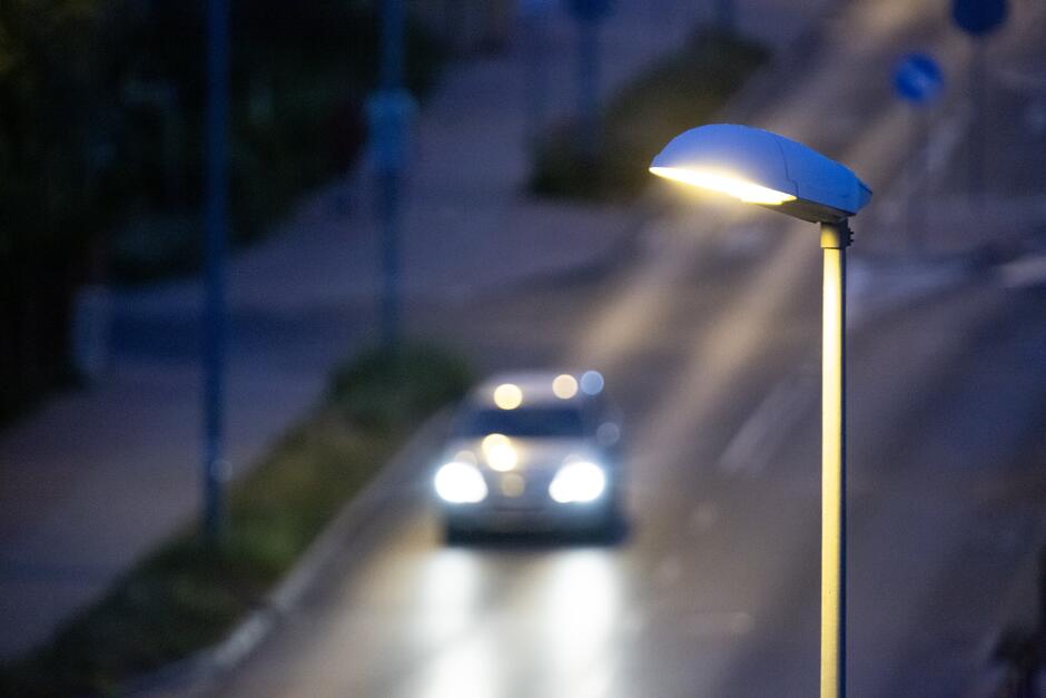 na zdjęciu świecąca latarnia, w tle samochód jadący z włączonymi światłami