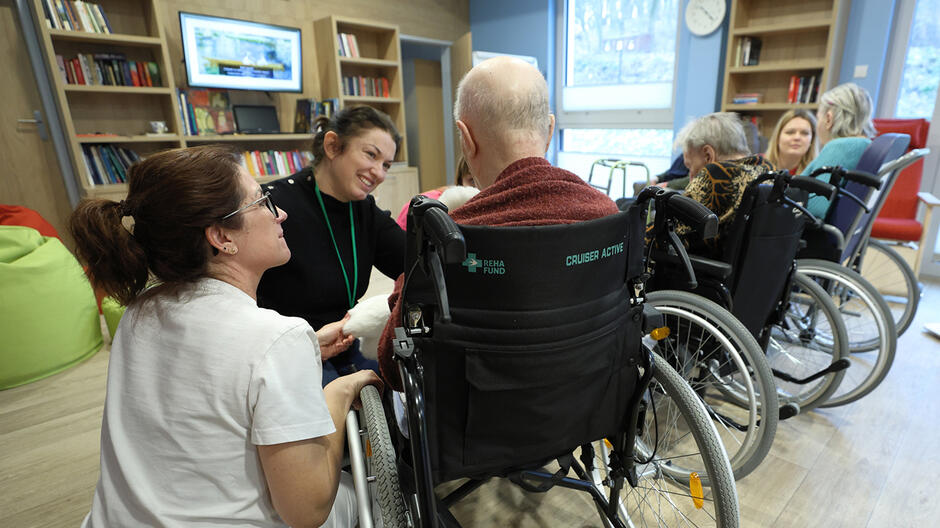 Trzy młode kobiety uśmiechają się do grupy starszych osób siedzących na wózkach inwalidzkich. Cała grupa siedzi w umeblowanej sali, przed nimi widoczny, wiszący na ścianie telewizor oraz regały wypełnione książkami
